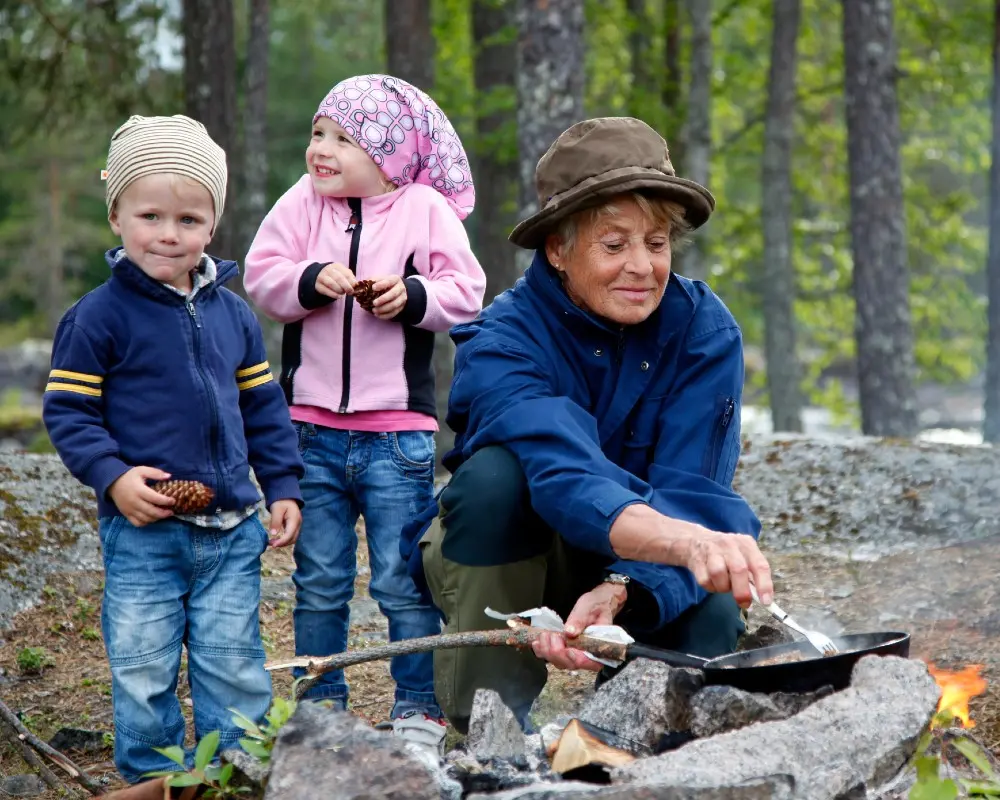 två små barn och en äldre dam i skogen. damen tänder en eld